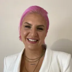 Porträt einer Frau mit brauner Haut und kurzen, pinkfarbenen Haaren, sie trägt einen weißen Blazer und lächelt strahlend.